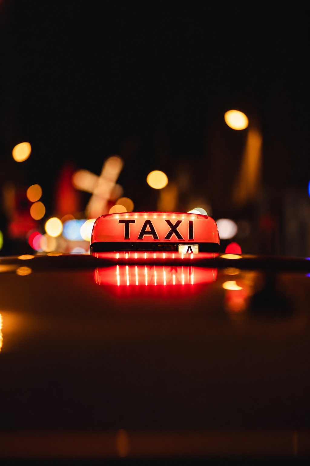 Un lumineux rouge TAXI posé sur le toit d’une voiture, éclairé de nuit devant des lumières de ville floues.